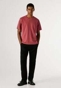 Bordeauxrode katoenen T-shirt met ronde hals en korte mouwen, gecombineerd met zwarte broek en zwarte schoenen, staand tegen een neutrale achtergrond.