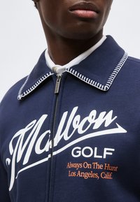 Gros plan d'une personne portant une veste de golf bleu marine zippée avec un col blanc cousu et le texte « Mullron Golf Always On The Hunt Los Angeles, Calif. »