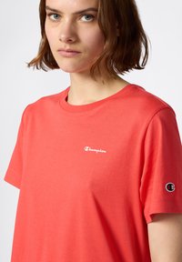 T-shirt w kolorze koralowym, wykonany z miękkiej bawełny, z okrągłym dekoltem, krótkimi rękawami oraz białym logo "Champion" na klatce piersiowej i małym logo na rękawie.