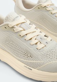 Chaussures de sport tricotées beige clair avec des lacets crème, semelle rembourrée étiquetée Corex Foam et nom de la marque Oysho sur la languette et le porte-lacets.