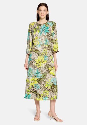 Kobieta w długiej sukience w kwiaty z niebieskimi, żółtymi i zielonymi liśćmi, stoi z rękami swobodnie opuszczonymi po bokach na białym tle.