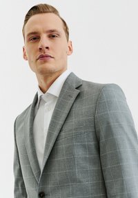 Szary, w kratę marynarka z wyciętym kołnierzem, pojedynczym zapięciem na guzik i naszytą kieszenią. Nosi się ją na białej koszuli z guzikami. Gładka tekstura.