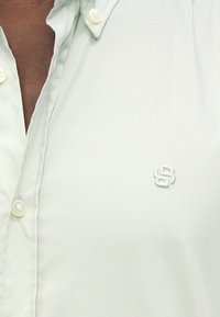 Ανοιχτό πράσινο πουκάμισο με κουμπιά, φτιαγμένο από μαλακό ύφασμα, με διακριτικό κεντημένο λογότυπο και λευκά πλαστικά κουμπιά.