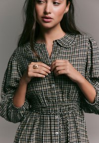 Robe à carreaux marron et blanc avec un col, une taille froncée et des manches bouffantes. Elle présente des boutons sur le devant et un tissu texturé.