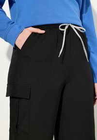Zwarte cargobroek met een zilveren trekkoord, voorzien van twee zijzakken en een gladde textuur. Gekocht met een blauwe lange mouwen top.