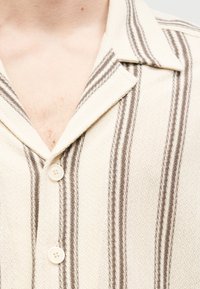 Kortærmet skjorte med cremefarvet bund og brune lodrette striber, med krave og to knapper lavet af et glat materiale.