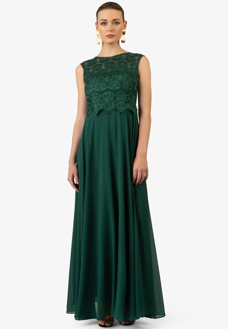 Lang groene jurk tot de vloer met een kanten top en een golvende rand, en een gladde, vloeiende rok. Aangevuld met minimale accessoires.