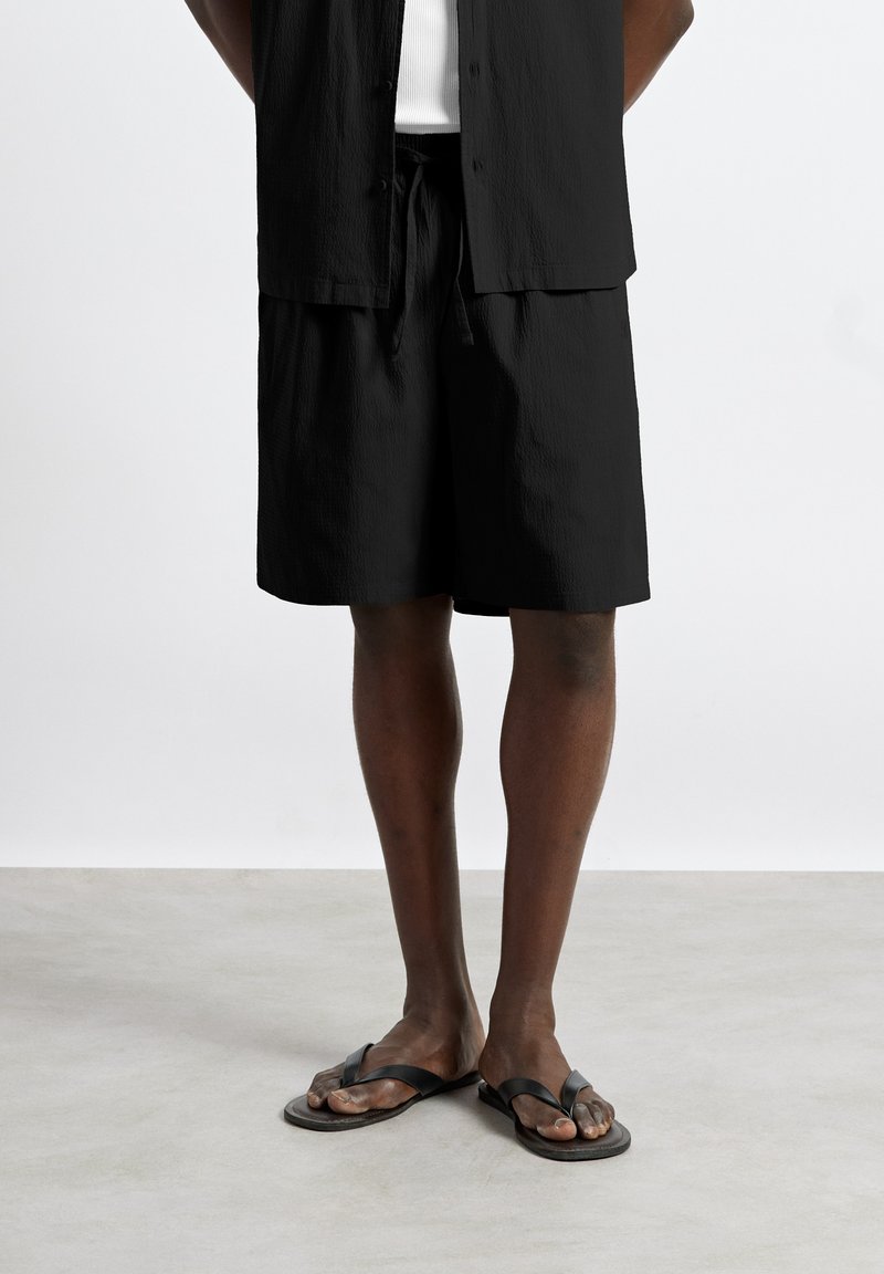 Personne portant un short noir, une chemise noire, un sous-vêtement blanc et des sandales noires, debout sur un sol gris clair devant un fond blanc.
