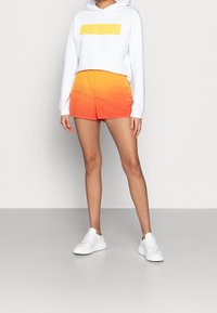 Vit croppad huvtröja med gult logotyp, i kombination med orange gradient-short. Inkluderar också vita sneakers, på en neutral bakgrund.
