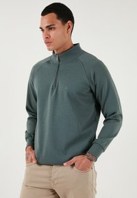 Sudadera de media cremallera verde hecha de tejido texturizado. Presenta un alto cuello, puños acanalados y un pequeño logotipo en la parte delantera.