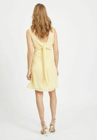 Vestido amarillo sin mangas con un diseño de atado en la parte de atrás, de longitud hasta la rodilla, hecho de tejido ligero. Presenta un escote en V profundo y un brillo sutil.