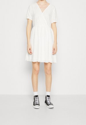 Kvinde iført en kort hvid øjlet kjole med korte ærmer og V-hals, kombineret med hvide sokker og sorte high-top sneakers.