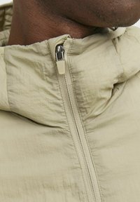 Veste zippée beige clair en tissu texturé avec capuche. Elle présente une fermeture éclair contrastante et une coupe décontractée au niveau du cou et des épaules.