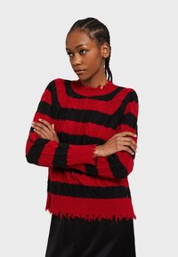 Sveter s červenými a čiernymi pruhmi, pletený s textúrovaným povrchom, má okrúhly výstrih a strapcovú spodnú časť. Obsahuje dlhé rukávy a voľný strih.