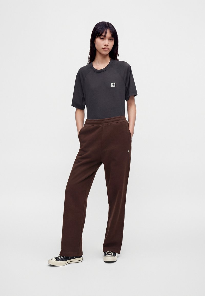 Junge Frau steht mit den Händen in den Taschen, trägt ein schwarzes T-Shirt, braune lockere Hose und schwarze flache Sneakers vor weißem Hintergrund.