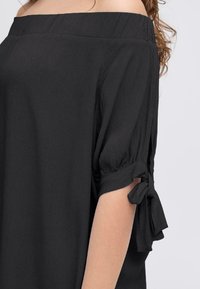 Top nero scollato con spalle scoperte e maniche a palloncino corte, con dettagli legati ai polsini. Il tessuto sembra leggero e leggermente strutturato.