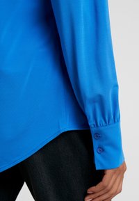 Blå långärmad skjorta med en slät textur, med knappade manschetter och en böjd nederkant. Matchas med mörk denim.