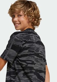 Camisa de patrón de camuflaje negro y gris con mangas cortas, que cuenta con un cuello redondo acanalado y tres rayas verticales en el hombro.