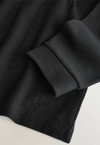 Schwarzes Langarmshirt aus strukturierter Stoff, mit einem dezenten geometrischen Muster an der Seite und einem gerippten Bündchen für zusätzliche Details.