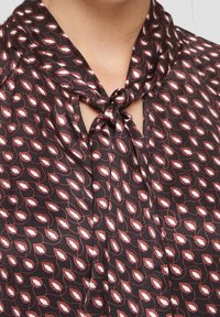 Bluse aus dunklem Stoff mit rot-weißer Blätteroptik, versehen mit einem gebundenen Kragen und einer Schlüssellochöffnung am Hals. Seidige Textur.