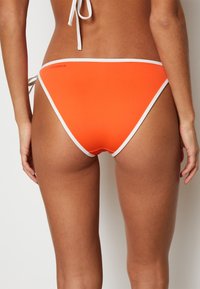 Klarorange bikiniunderdel med vit kant, som har en sömlös design och justerbara knytband för en anpassad passform. Slät tygtextur.