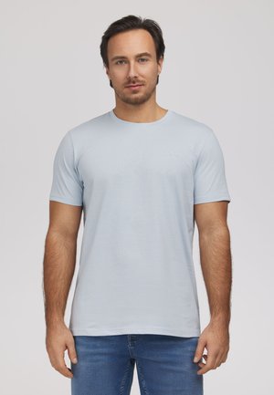 Mężczyzna z krótkimi ciemnymi włosami, ubrany w jasnoniebieski t-shirt z krótkim rękawem i niebieskie dżinsy, stoi na tle jednolitego jasnego tła.