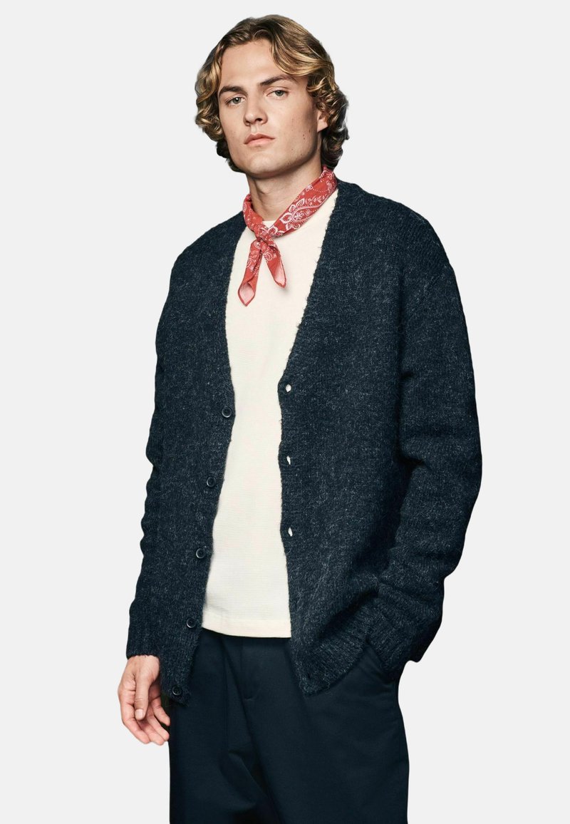Dunkelgrauer gestrickter Cardigan mit strukturiertem Finish, ausgestattet mit einem V-Ausschnitt, Knopfleiste und langen Ärmeln, getragen über einem weißen Hemd.