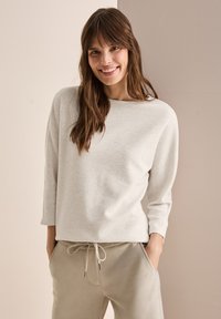 Helle beige, ribbelte Strickjacke mit weitem Ausschnitt, langen Ärmeln und lockerer Passform. Kombiniert mit hellgrauen Jogginghosen mit einem Kordelzugbund.