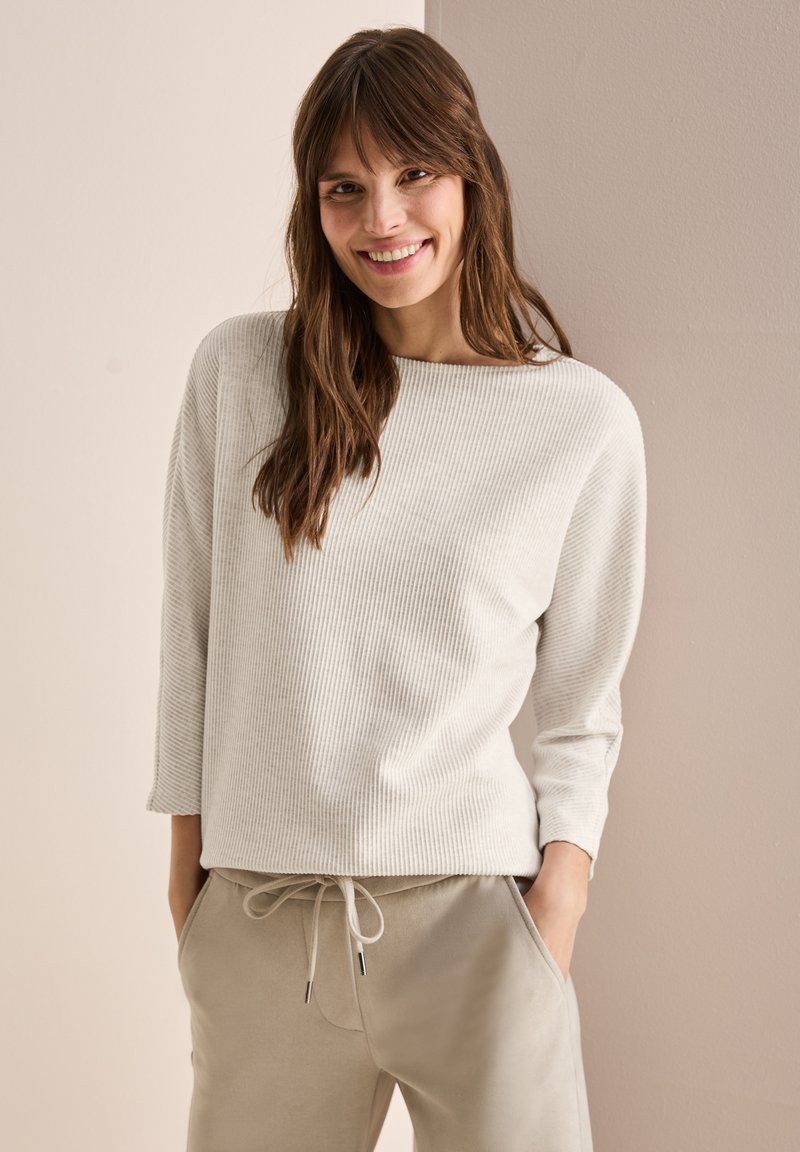 Helle beige, ribbelte Strickjacke mit weitem Ausschnitt, langen Ärmeln und lockerer Passform. Kombiniert mit hellgrauen Jogginghosen mit einem Kordelzugbund.