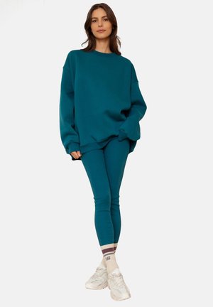 Turkusowy, oversize'owy sweter i dopasowane legginsy o miękkiej teksturze. Model nosi białe sneakersy z beżowymi i burgundowymi paskami na skarpetkach.
