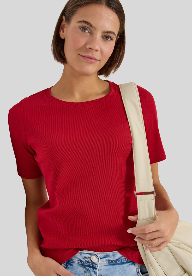 Femme aux cheveux bruns courts portant un t-shirt rouge à manches courtes, un jean bleu clair et tenant une sangle de sac beige sur son épaule.