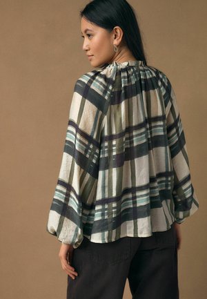 Sieviete ar gariem tumšiem matiem, tērpusies vaļīgā, garām piedurknēm klātā pleda blūzē zaļas, melnas un baltas krāsas toņos, valkā tumšas bikses un atrodas priekšā brūnam fonam.