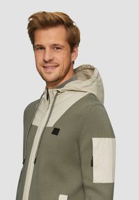 Khaki Kapuzenpullover aus strukturiertem Stoff, mit cremefarbenen Akzenten, Reißverschlusstaschen und einer Kapuze mit Kordelzug. Schwarzes Logo auf der Vorderseite.