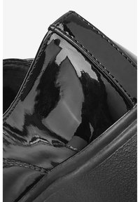 Chaussure en cuir verni noir avec une finition brillante, bout arrondi et détail de couture le long de la tige. La semelle présente une base en caoutchouc texturé.