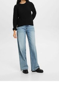 Czarny sweter z dzianiny z okrągłym dekoltem, zestawiony z jasnoniebieskimi spodniami o szerokich nogawkach. Model nosi czarne buty na platformie.
