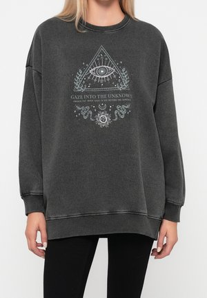 Kobieta ubrana w ciemnoszarą, oversize'ową bluzę z mistycznym okiem wewnątrz trójkąta, wężem, słońcem, księżycem, roślinami oraz napisem "Patrz w nieznane."
