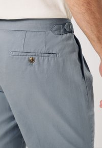Pantaloni su misura color azzurro chiaro con tasca posteriore con dettaglio bottone, passanti per cintura e una texture liscia. Tasche laterali incluse.