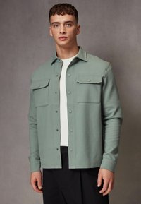 Chaqueta de camisa estructurada verde claro, confeccionada en una mezcla de algodón, con dos bolsillos en el pecho y cierre de botones a presión, llevada sobre una camiseta blanca.