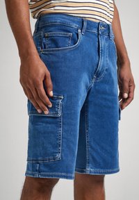 Jeanskorta shorts i medelblått med slät textur, fem fickor inklusive en sidosnickepocket, och synliga sömnadsdetaljer.