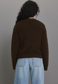 Tumepruun kudumissweater, millel on sooniline tekstuur ja pikad varrukad, koos helesiniste laia säärega teksadega.