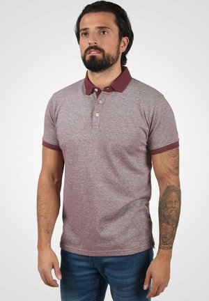 Bordeaux polo-shirt van een gestructureerde stof, met een knoopsluiting van twee knopen en contrasterende donkere kraag- en mouwafwerking. Bevat een tattoo op de onderarm.