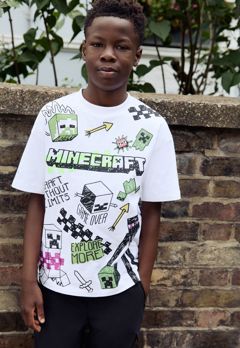 Garçon debout à l'extérieur devant un mur en briques, portant un t-shirt blanc Minecraft avec des personnages et des graphismes du jeu.