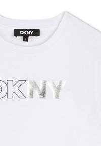 T-shirt en coton blanc arborant un logo métallique noir et argent "DKNY". Encolure ras du cou classique, texture lisse, manches courtes.