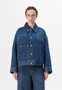 Džínová bunda v tmavě modré barvě, s dvěma předními kapsami, stříbrnými cvočky na límci a uvolněným, oversize střihem.