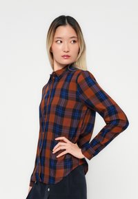 Plaid button-up shirt in brown, blue, and black tones. Features a collar, long sleeves, and a curved hem. Made from a smooth fabric.