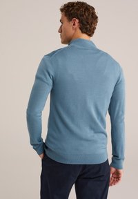 Mees, kellel on seljas helesinine pikkade varrukatega kampsun ja tumedad püksid, seisab käed taskutes, pööratud eraldi tausta poole.