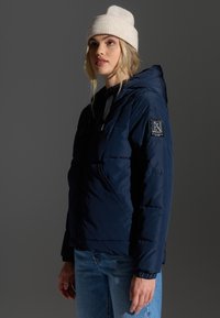 Mørkeblå polstret jakke med hætte, prydet med et logo- patch på ærmet, parret med lyseblå jeans og en cremefarvet strikket hue.
