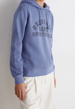 Personne portant un sweat à capuche bleu clair avec le texte "Original GANT Sportswear", associé à un pantalon blanc ample, sur un fond uni.