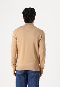 Beżowy dzianinowy sweter z długimi rękawami i wysokim kołnierzem, z prążkowanymi mankietami i dołem. Nosi się go na ciemnoniebieskich dżinsach.