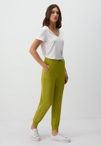 Grüne Jogginghosen mit Seitentaschen und elastischen Bündchen, kombiniert mit einem weißen V-Ausschnitt-T-Shirt. Modell trägt weiße Turnschuhe. Minimalistisches Design.
